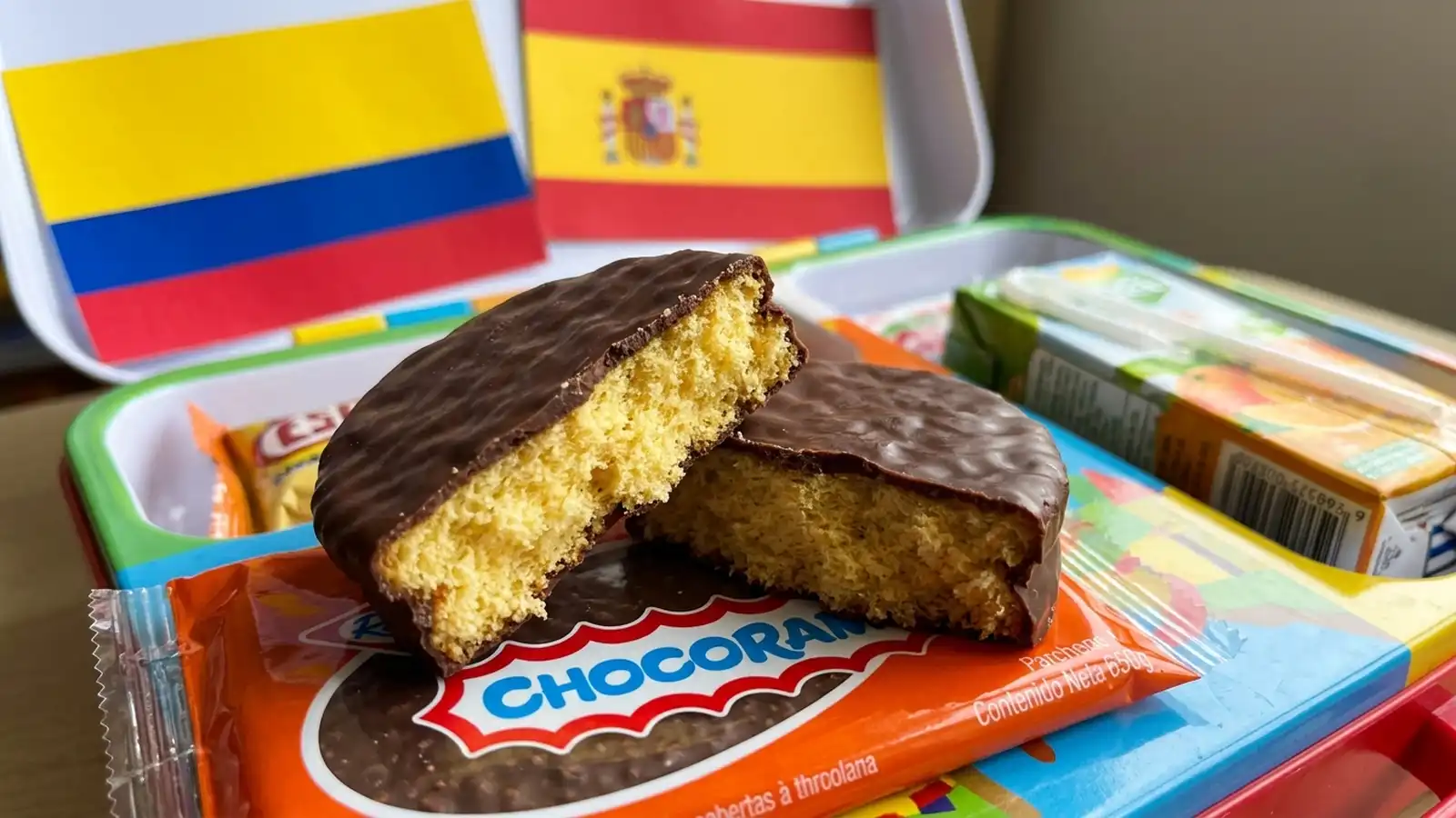 Un Chocoramo partido por la mitad mostrando su bizcocho amarillo y cubierta de chocolate sobre su empaque naranja, situado dentro de una lonchera escolar con las banderas de Colombia y España de fondo.